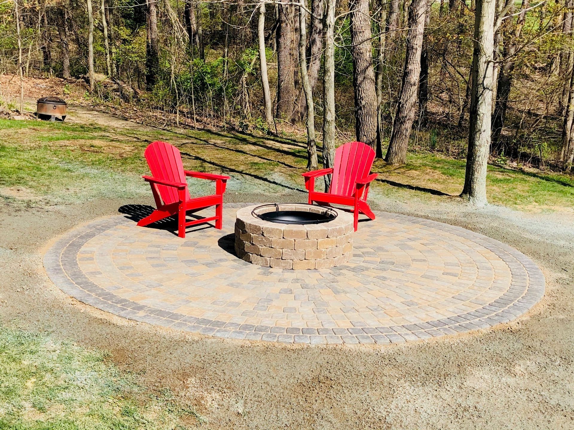 Two bright red Adirondack chairs face a circular stone fire pit on a paved patio in a wooded yard.