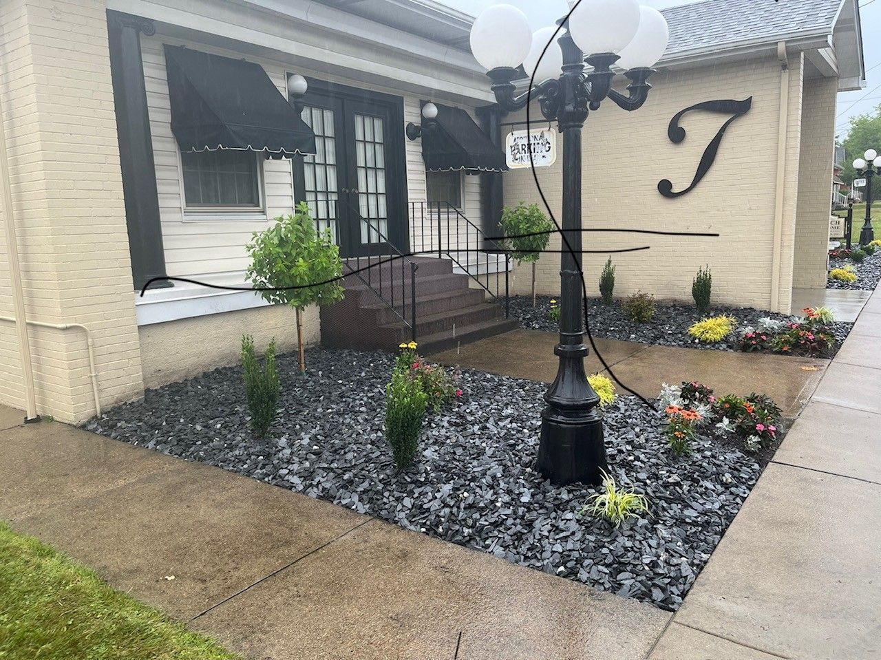 A building entrance with black awnings, dark stone landscaping, a streetlamp, and a large decorative letter 