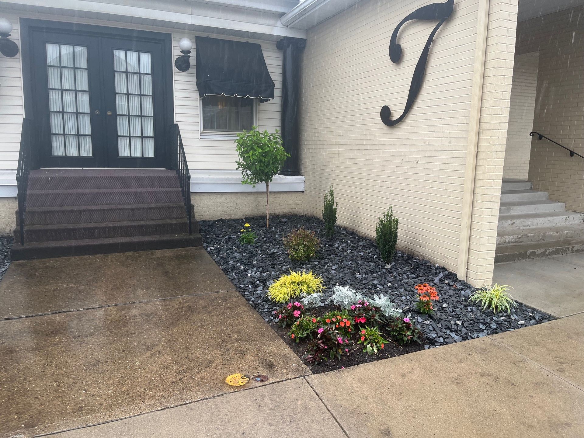 A building entrance with black double doors, stone steps, and a garden bed featuring grey rocks and colorful flowers.
