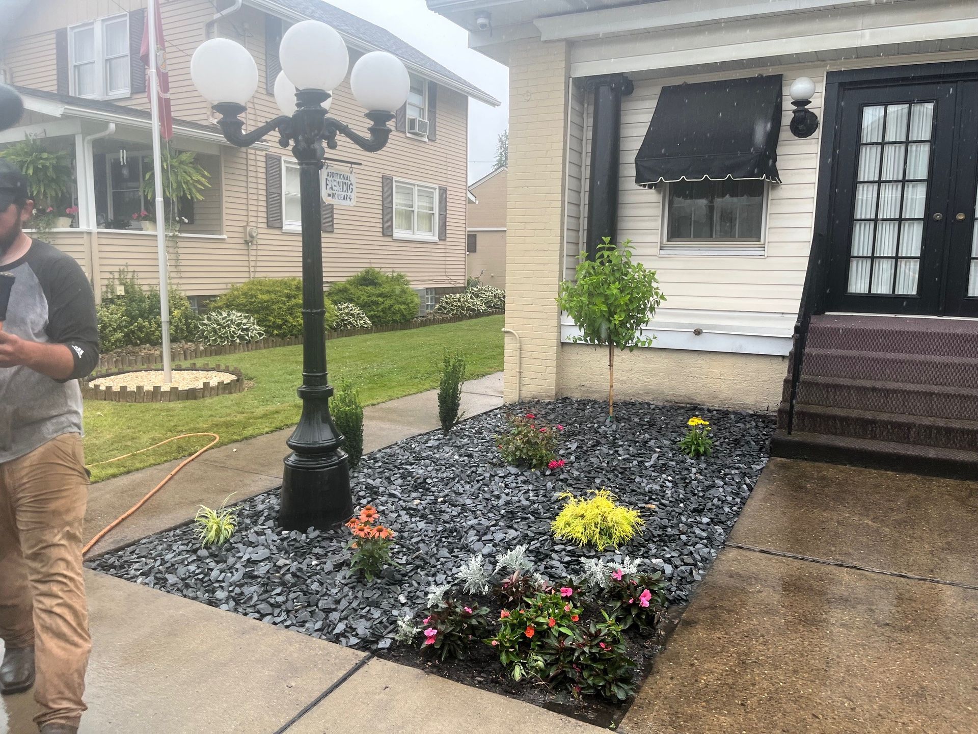 A person stands near a lamp post and a flower bed filled with dark grey mulch in front of a house with black doors.