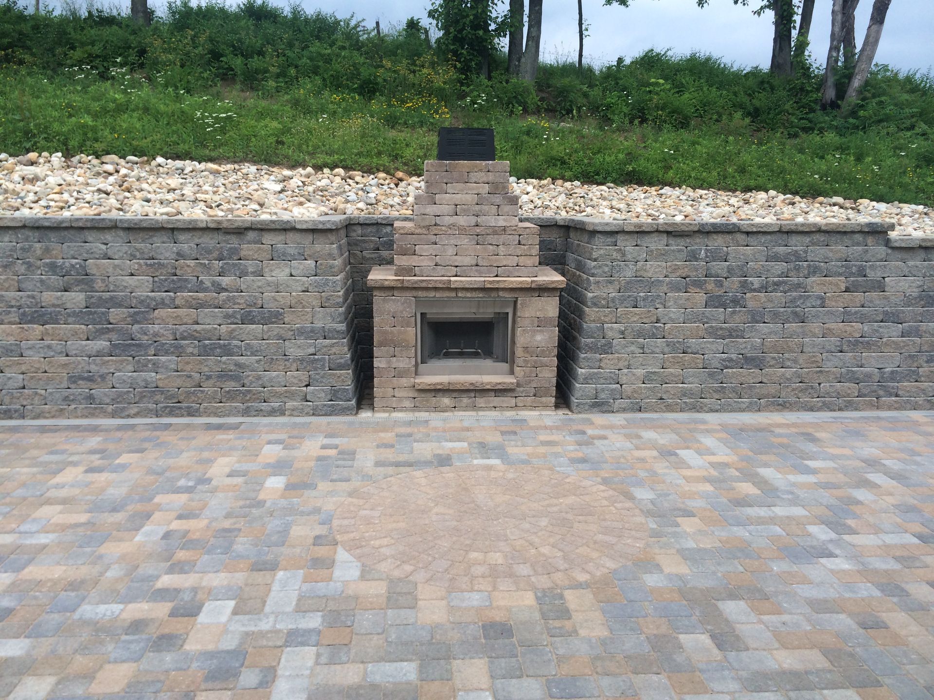An outdoor brick fireplace set into a multi-toned stone patio and retaining wall, backed by a grassy hill.