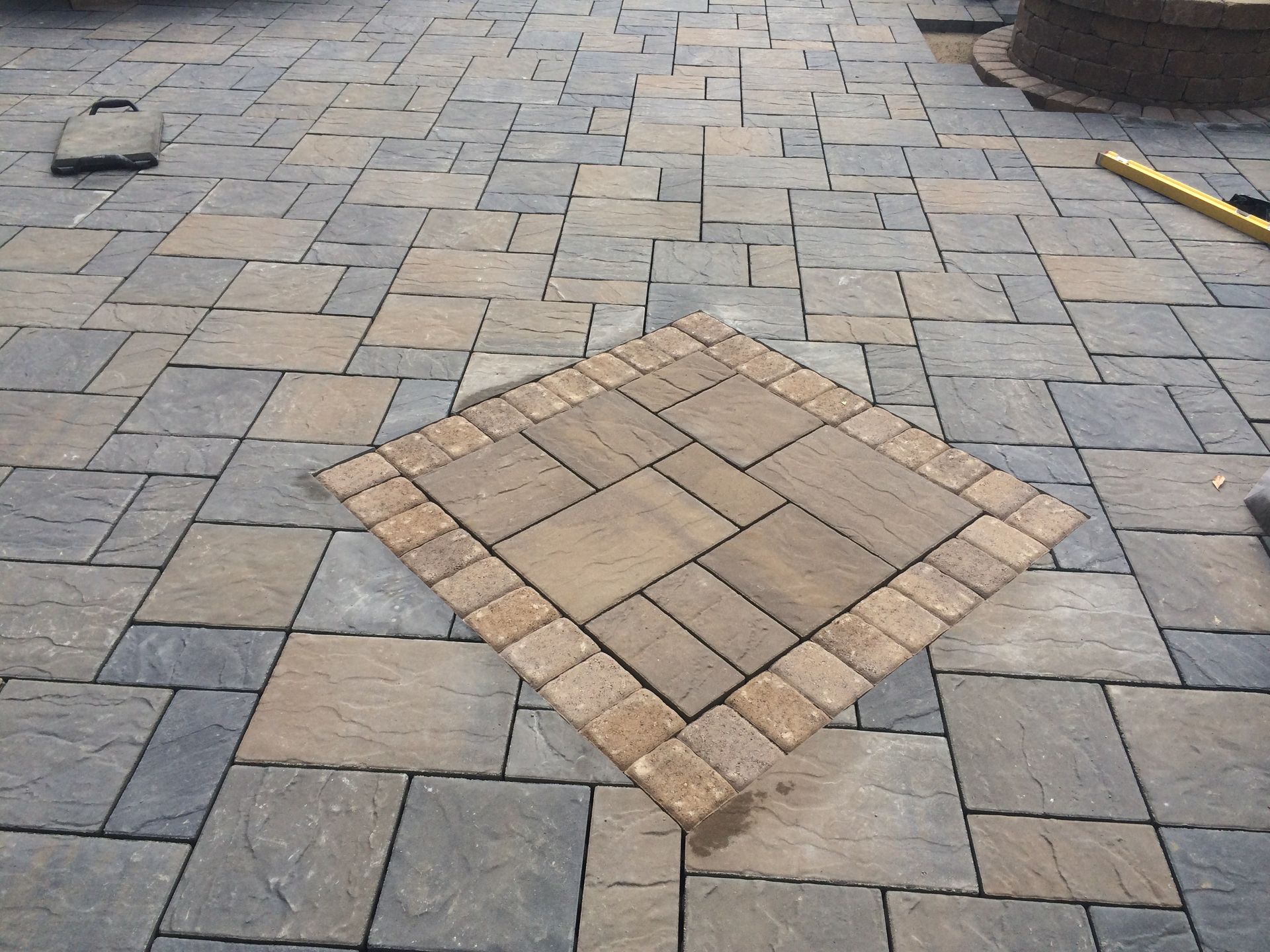 A stone patio with a textured, multi-colored paver pattern and a square central accent piece framed by a border of bricks.