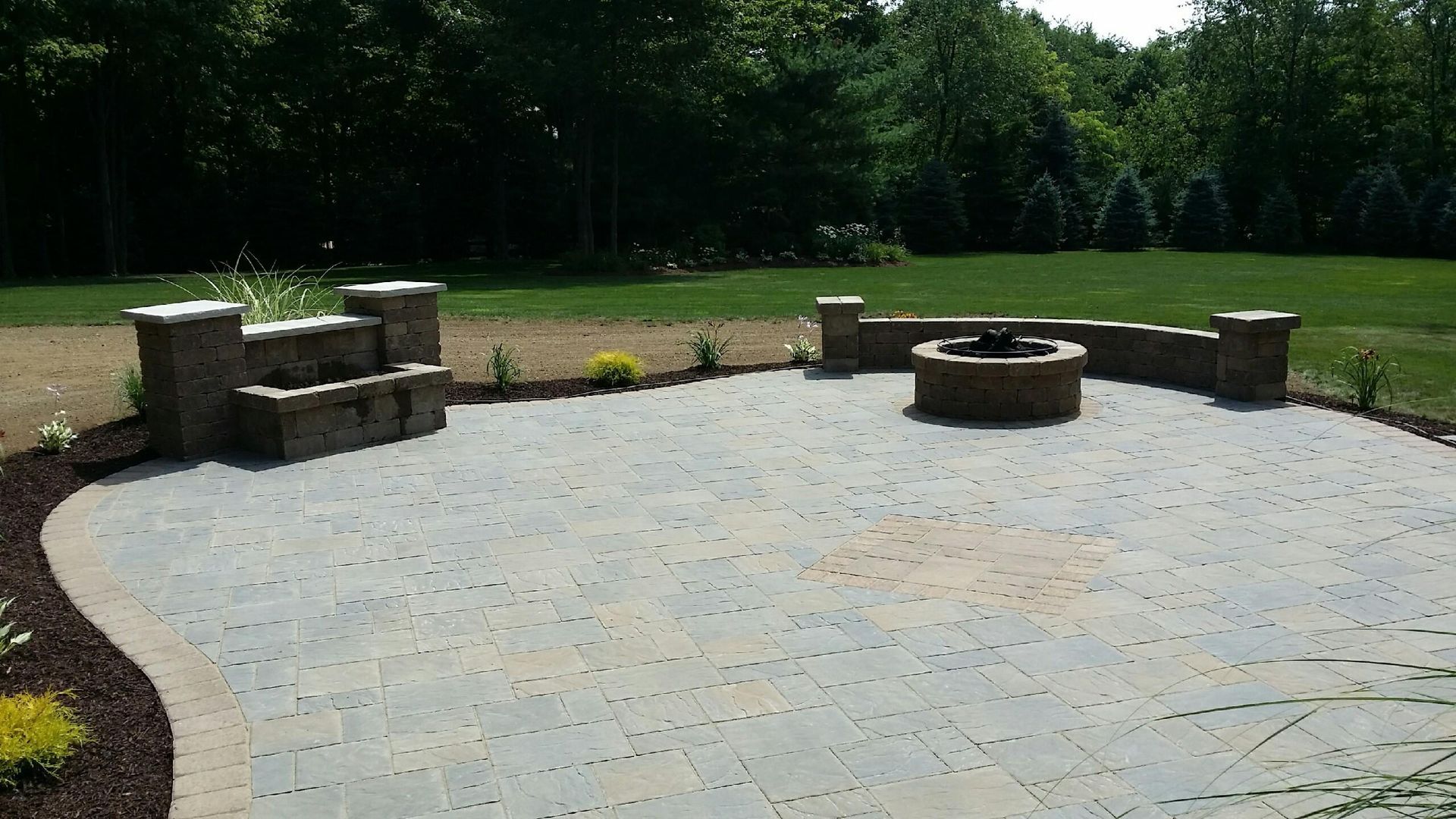 A circular stone patio features a built-in fire pit, stone seating wall, and pillars, set against a green lawn and trees.