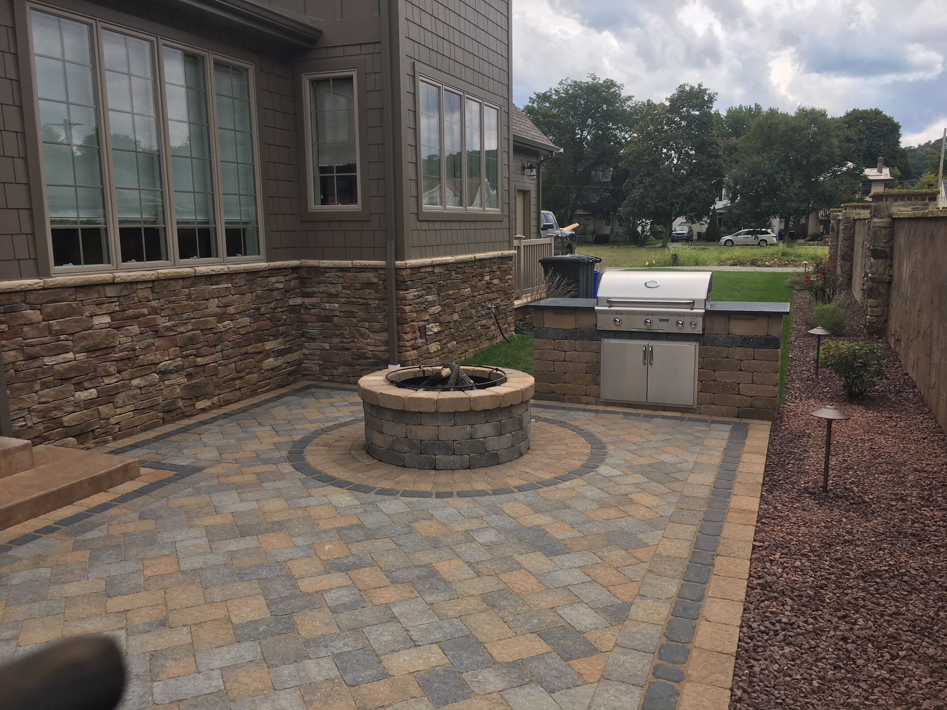 A stone fire pit sits in the center of a paved patio next to a house with a built-in outdoor grill and stone siding.