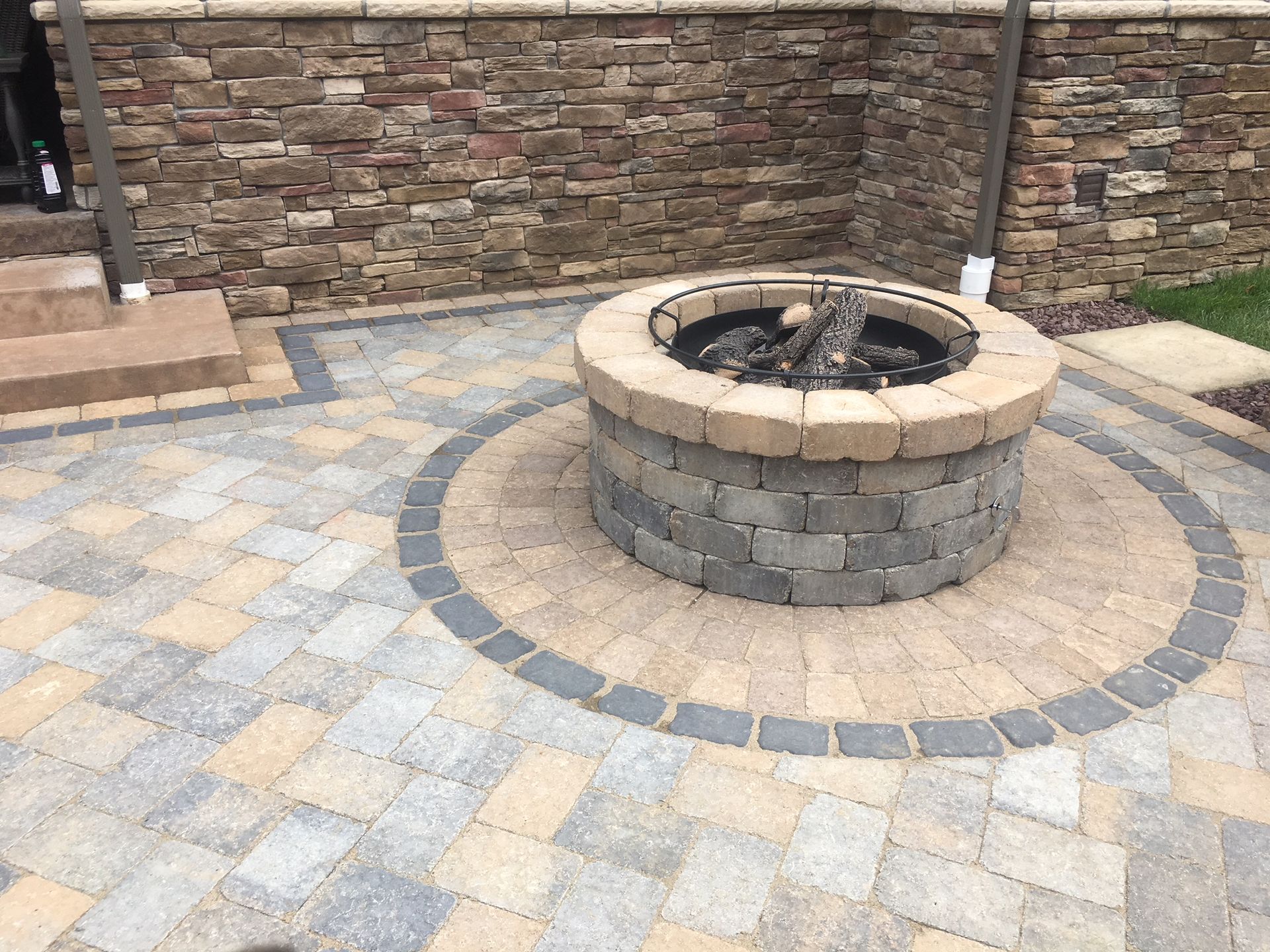 A stone fire pit centered on a multi-colored paver patio next to a stone-clad house wall.