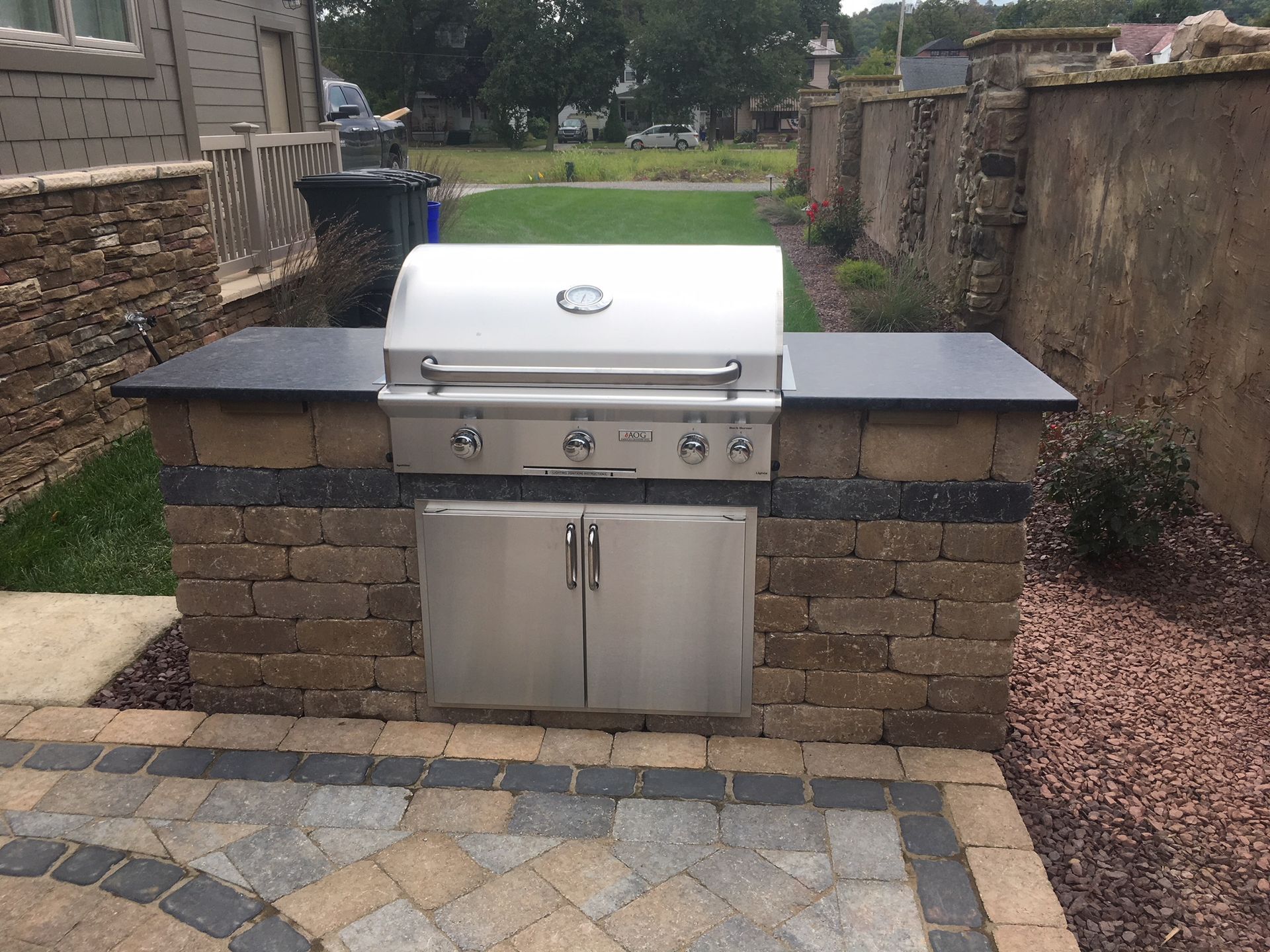 An outdoor stainless steel gas grill built into a stone masonry counter on a paved patio.