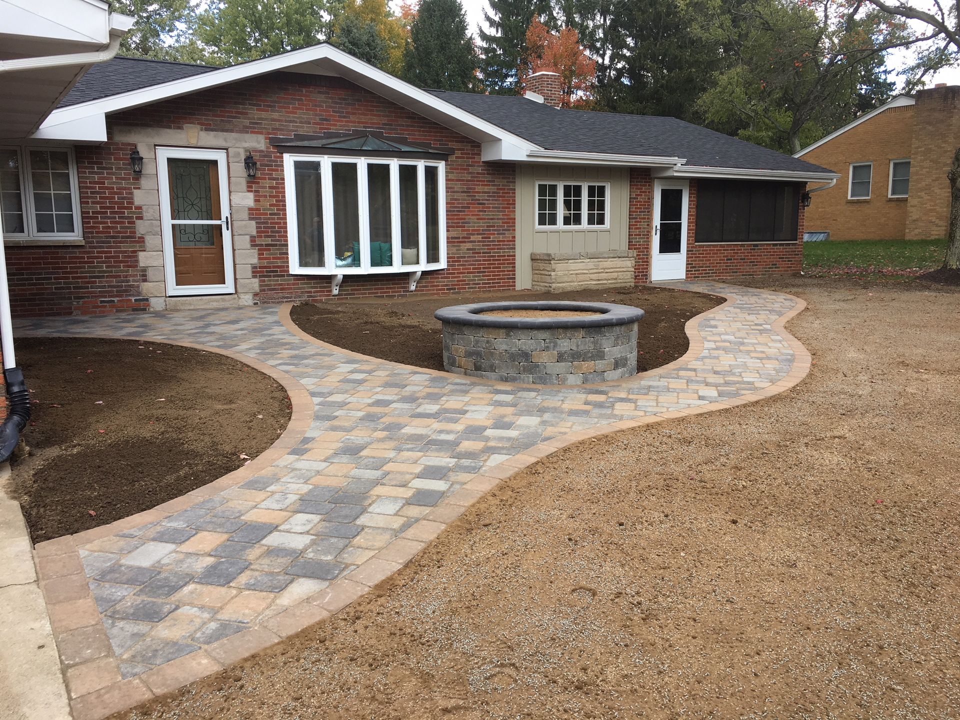 A winding paver walkway leading to a brick house with a central stone fire pit in the front yard.