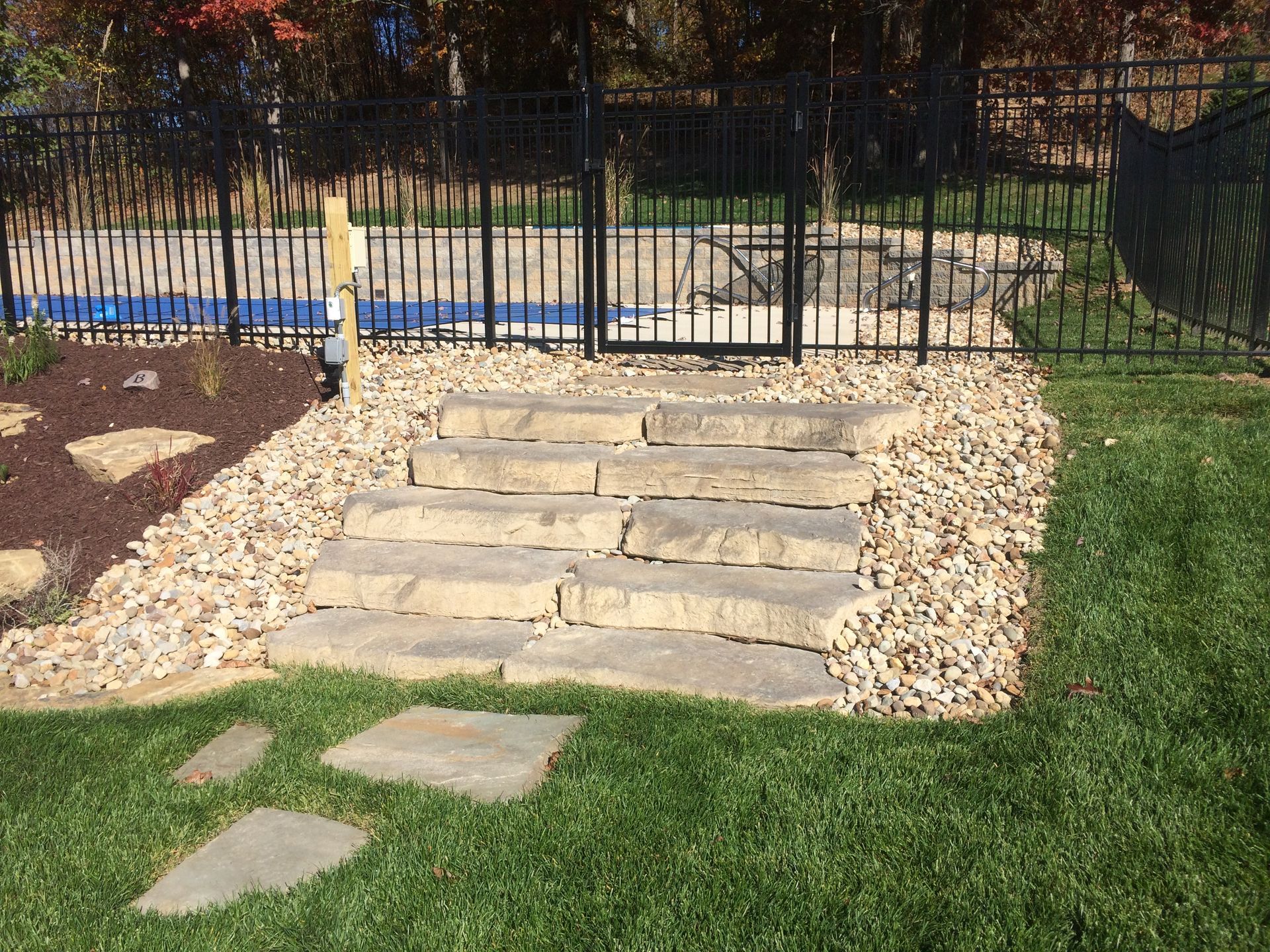 Natural stone steps lead through a gravel bed to a black metal fence and gate surrounding a backyard pool area.