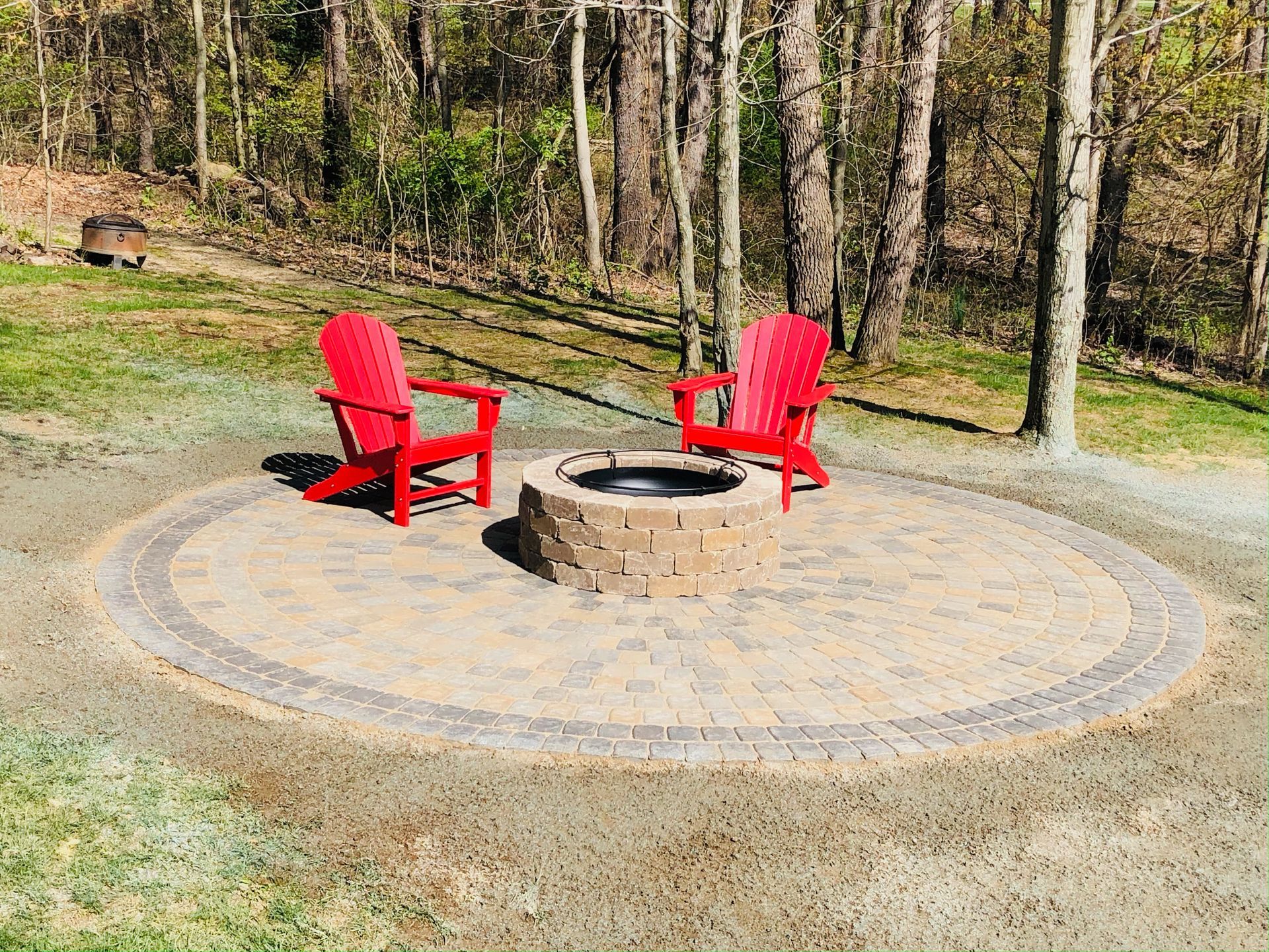 Two bright red Adirondack chairs sit on a circular stone patio around a circular stone fire pit in a wooded backyard.