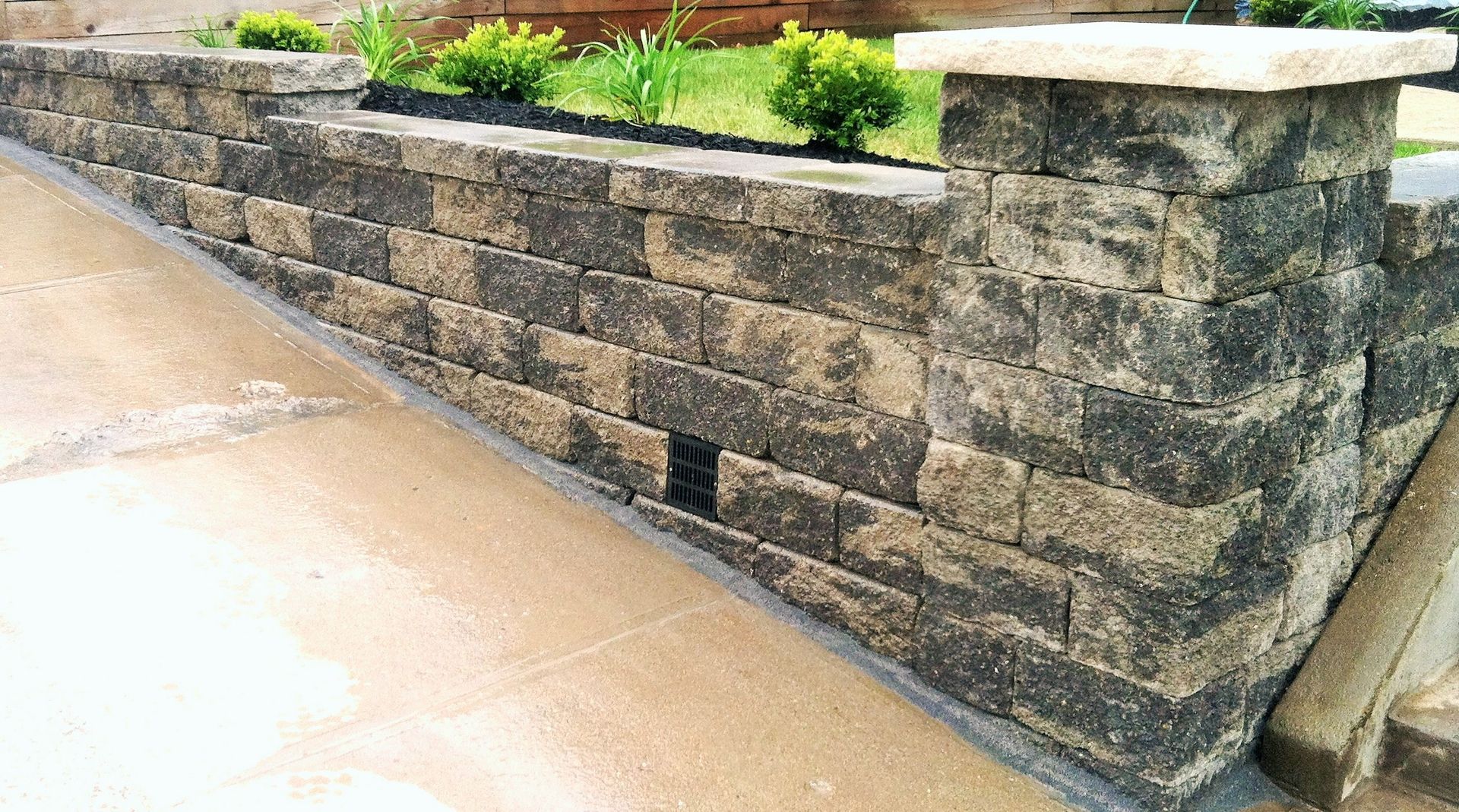 A low stone retaining wall with a square pillar and a small black vent, built along a concrete walkway.