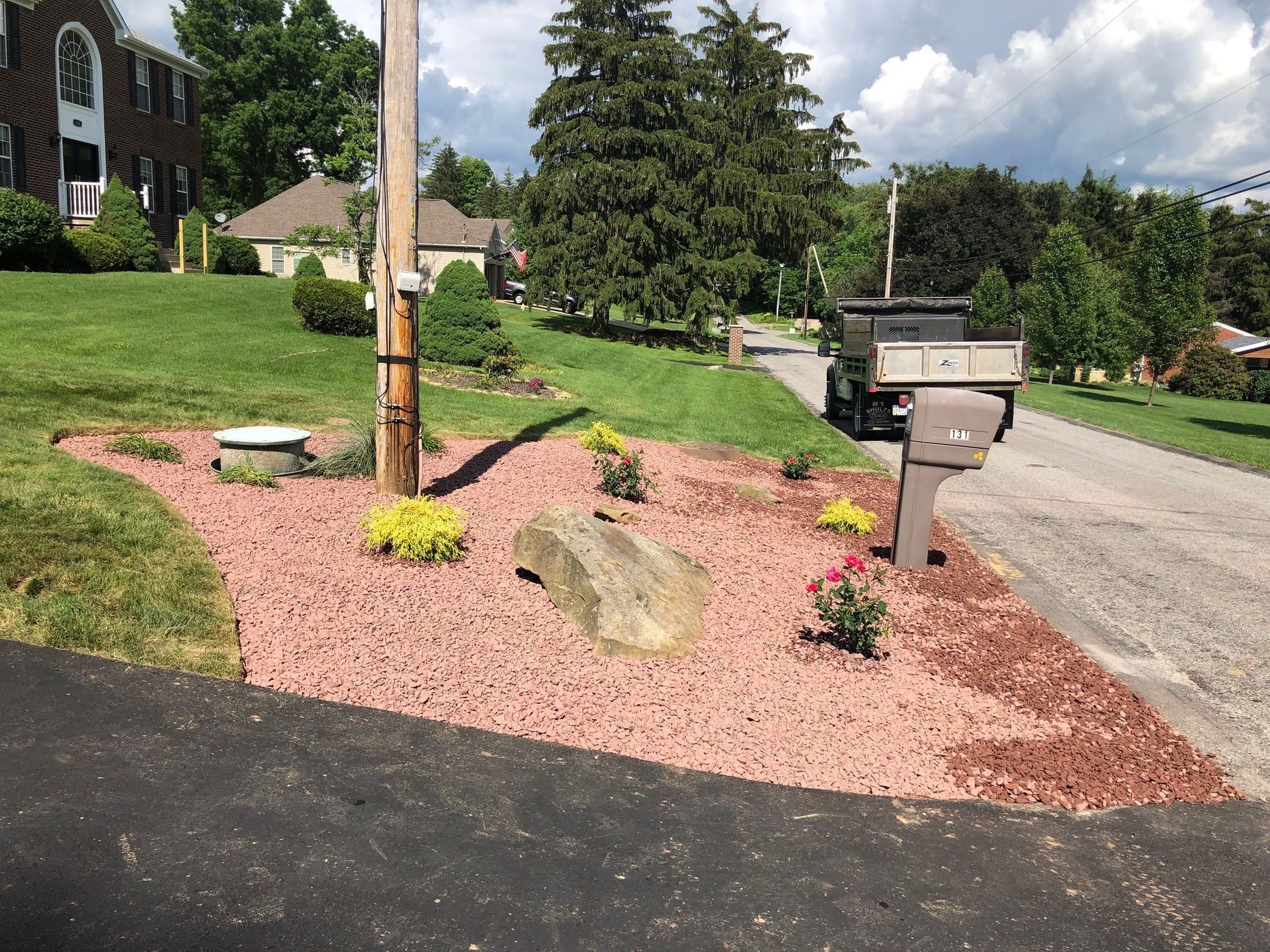 A landscaped front yard area featuring red mulch, a large decorative rock, small shrubs, and a mailbox by a paved driveway.