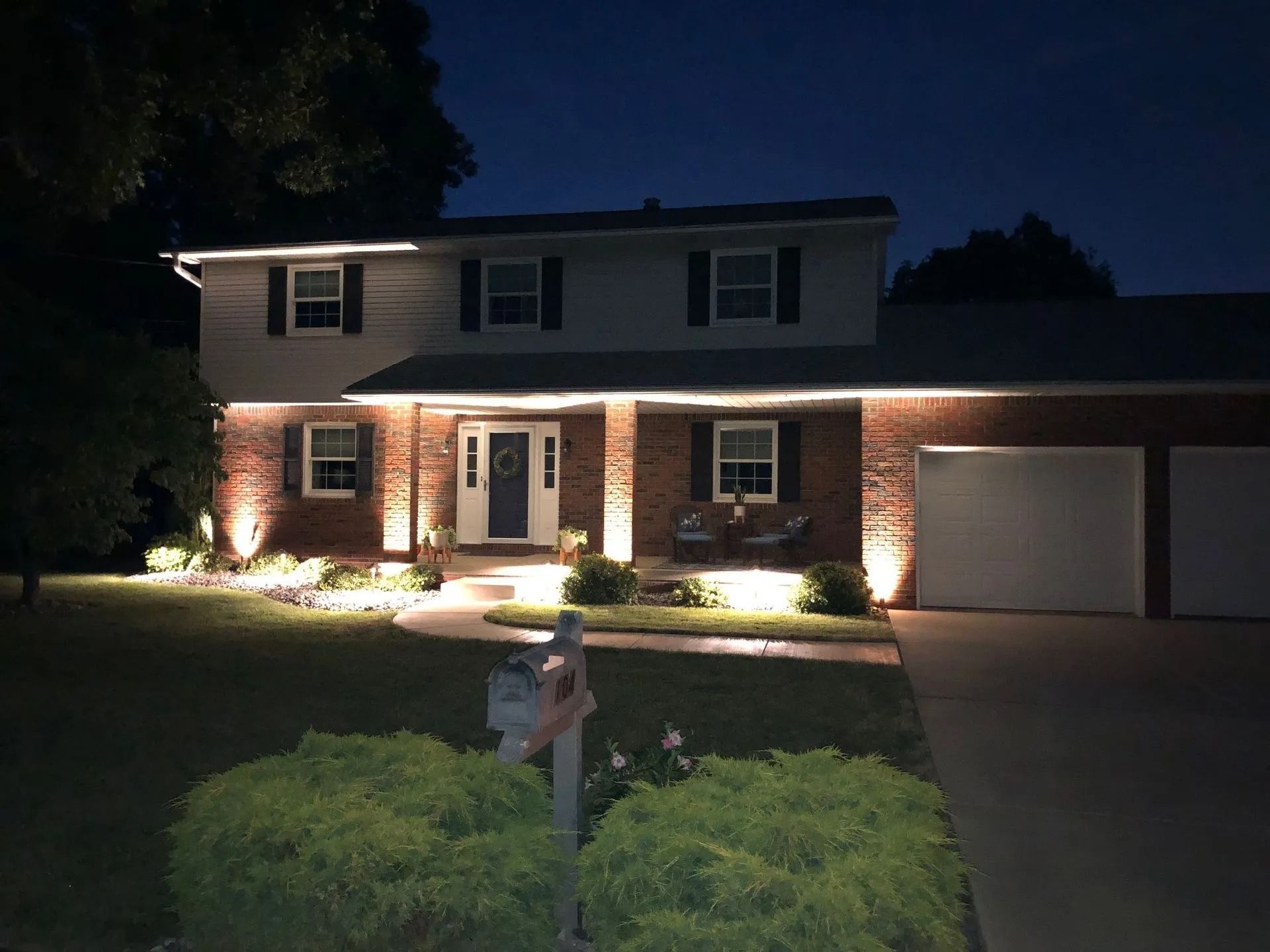 Lit up two-story house at night with lights outlining the roof, entrance, and landscaping.