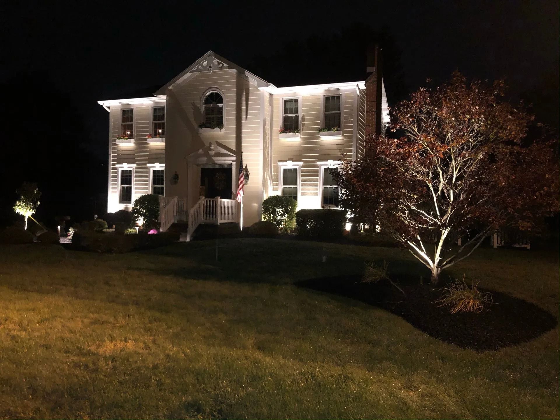 Two-story white house at night, lit with landscape lights. A tree with red leaves is illuminated in the foreground.