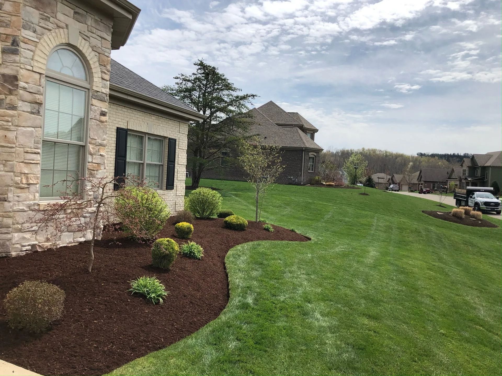 House with manicured lawn, mulch beds, and surrounding green space on a cloudy day.