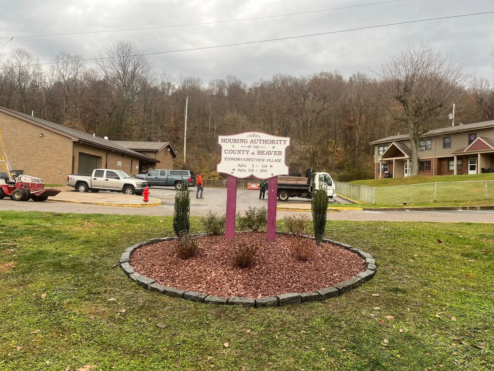 Sign for a housing community with vehicles and buildings in the background. Green grass and trees surround it.