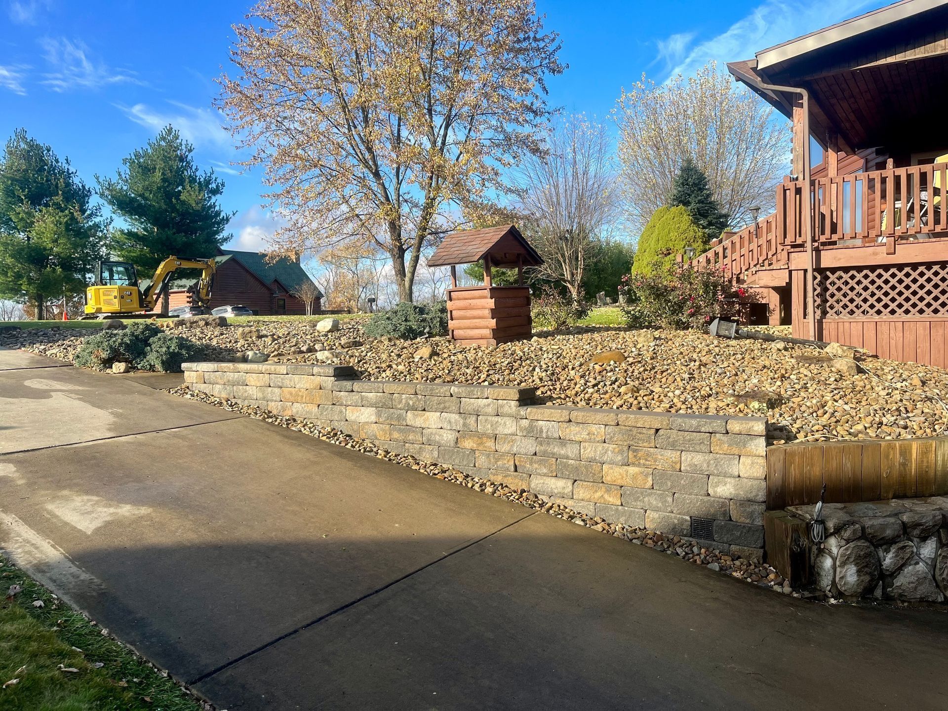 Asphalt driveway leading to a brown house with a well, rock retaining walls, and fall foliage under a blue sky.