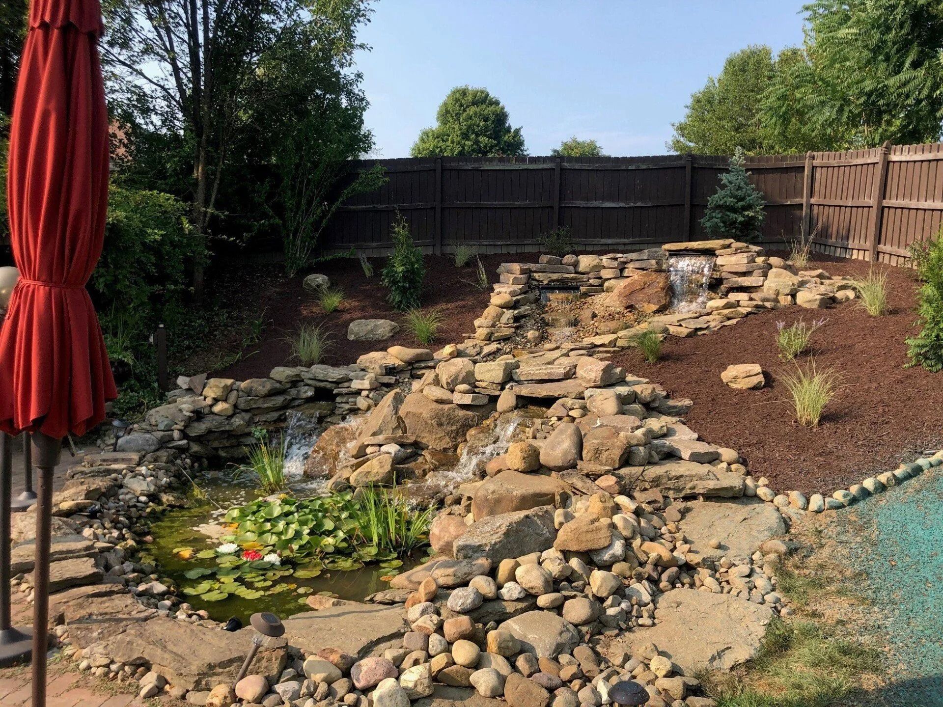Backyard water feature with cascading waterfall, pond with lily pads, rock edging, and a brown fence.