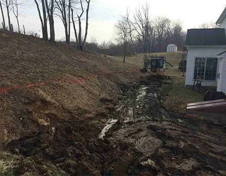 Muddy hillside with exposed earth, potential erosion. Tractor in distance, house on right.
