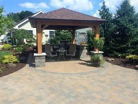 Outdoor gazebo with seating, surrounded by a paved patio and landscaping under a sunny sky.