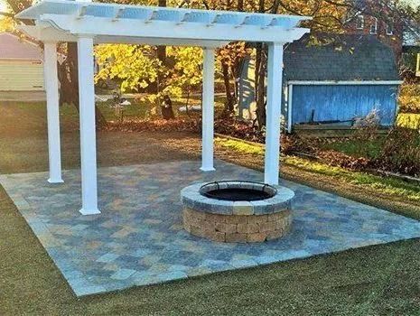 Pergola over a stone fire pit on a paved patio in a grassy yard, a blue shed in background.