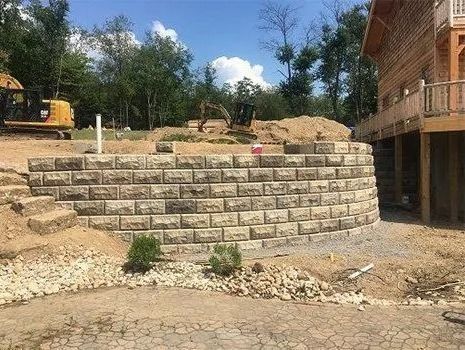 Curved stone retaining wall under construction near a wooden building, with machinery and dirt piles.