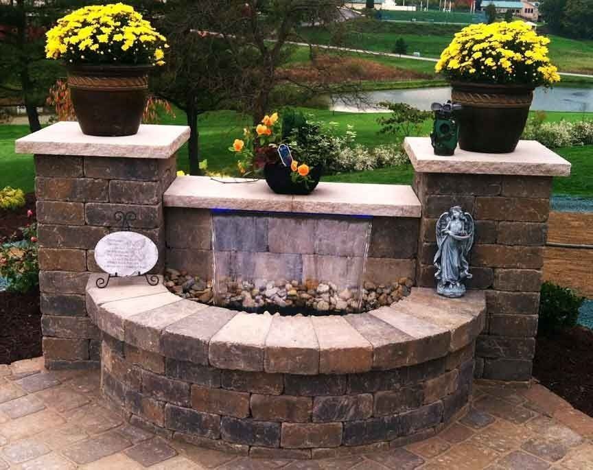 Stone water fountain with yellow mums in pots, a small waterfall, and a garden setting.