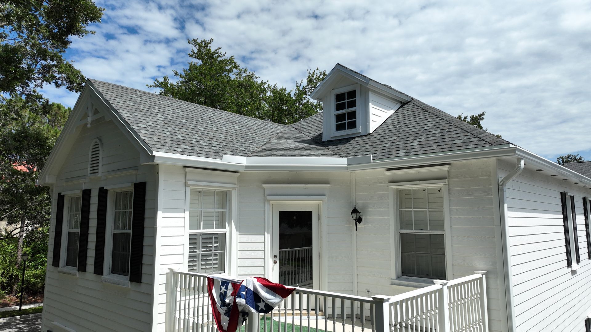  A small white home with a gray shingle roof, dormer window, porch railing, and patriotic bunting