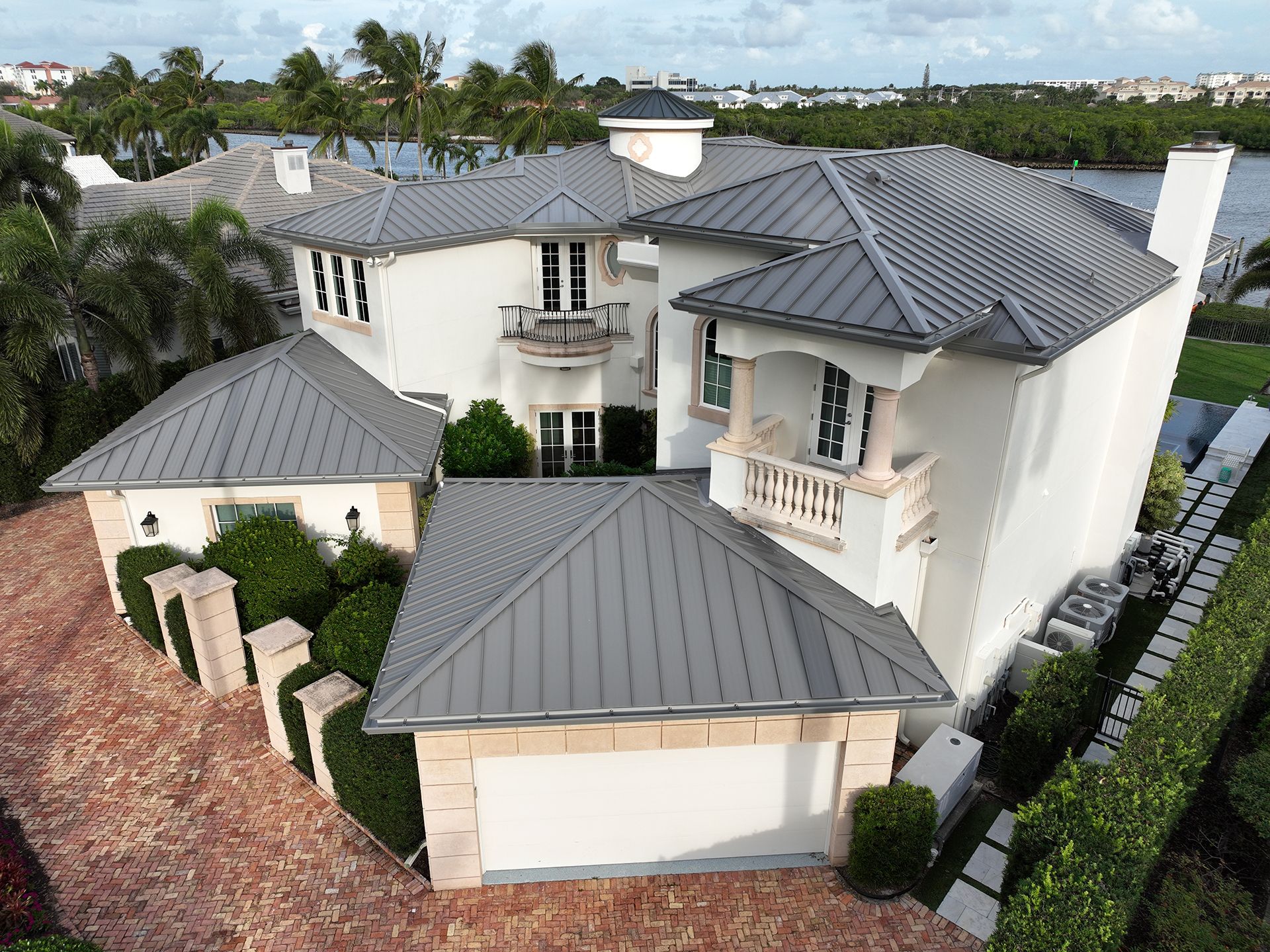 Large white house with gray metal roof, brick driveway, and lush landscaping near water.