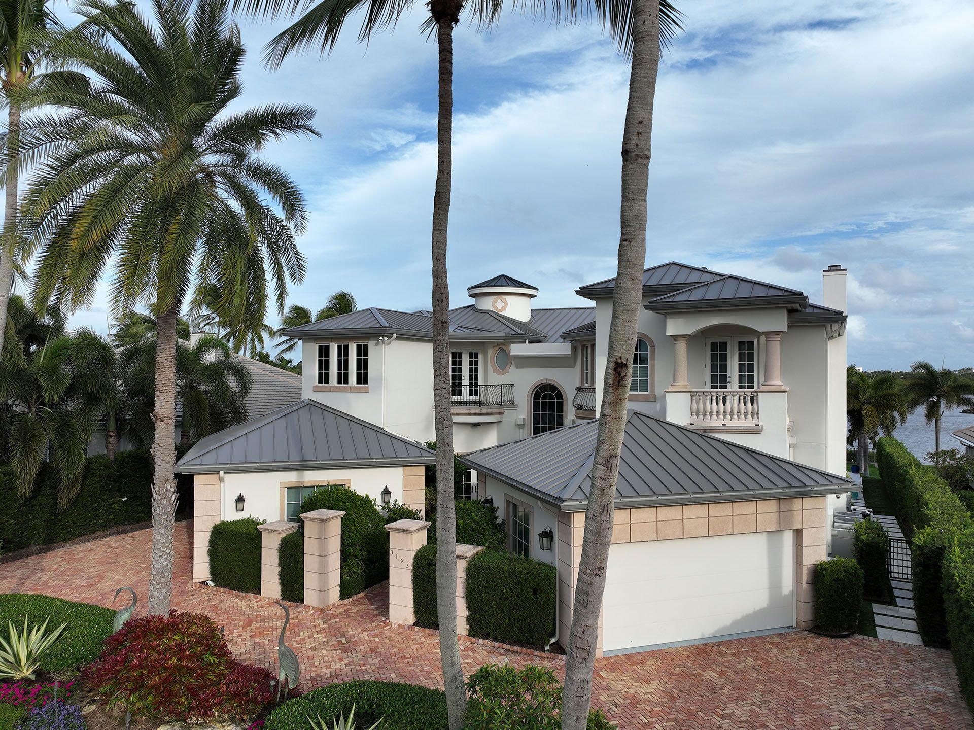 White stucco mansion with gray roof, palm trees, and brick driveway.