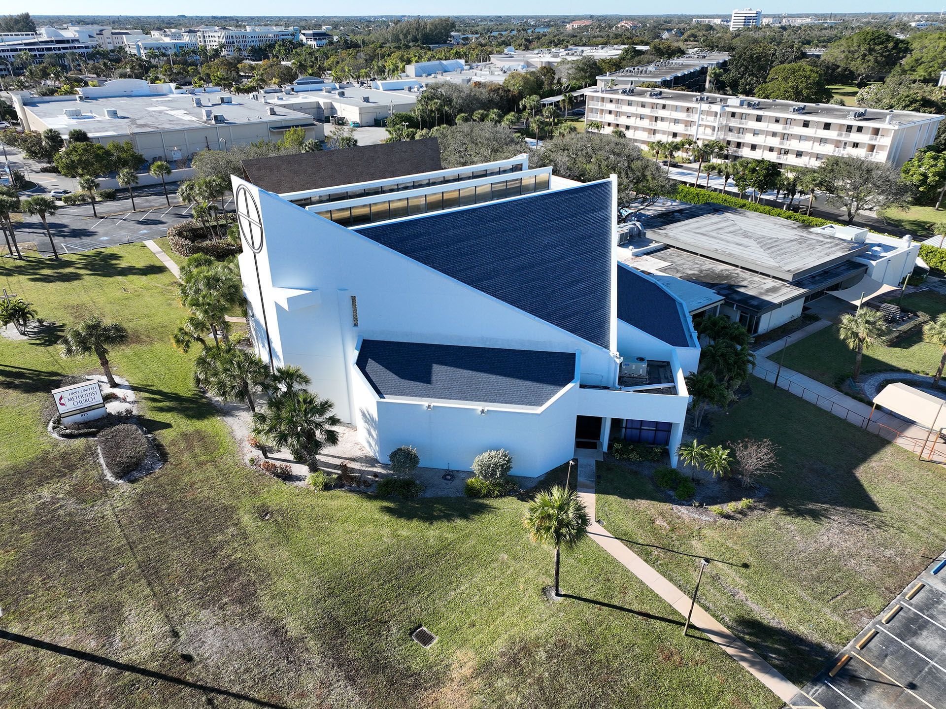 A modern white church building with a dark roof surrounded by buildings and trees on a sunny day.