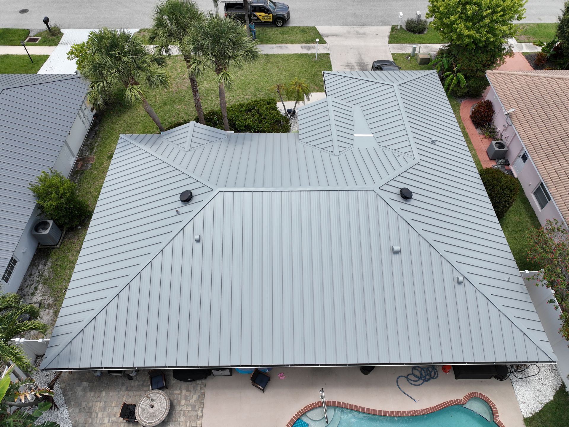 Aerial view of a gray metal roof on a house, with palm trees and a pool in the yard.