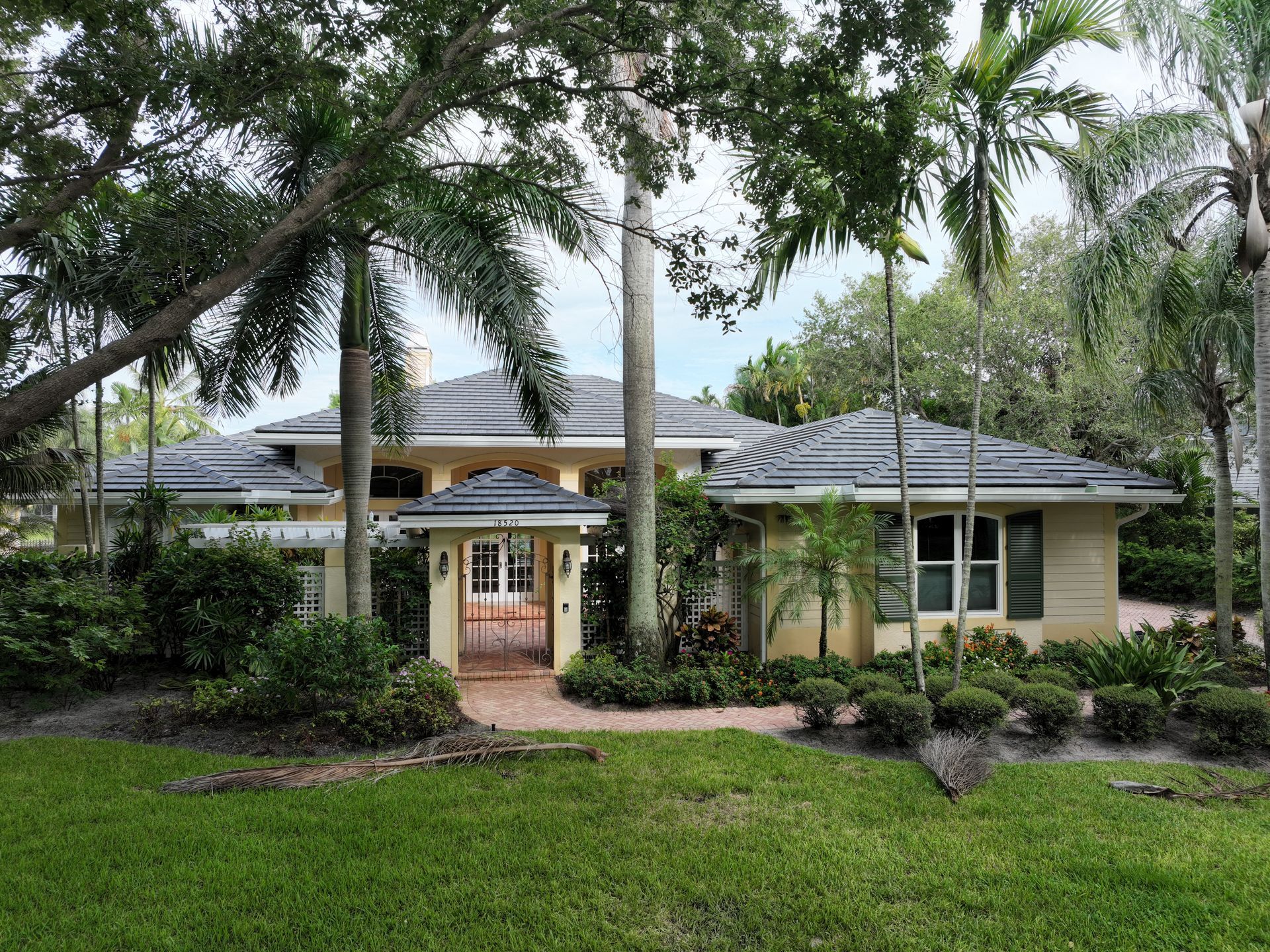 A large, light yellow house with a dark gray roof surrounded by lush greenery and palm trees.