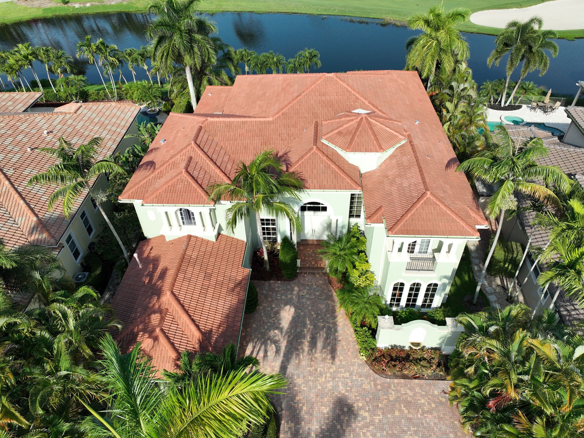 Overhead view of a light green house with a red tile roof. Palm trees surround the home near a body of water.