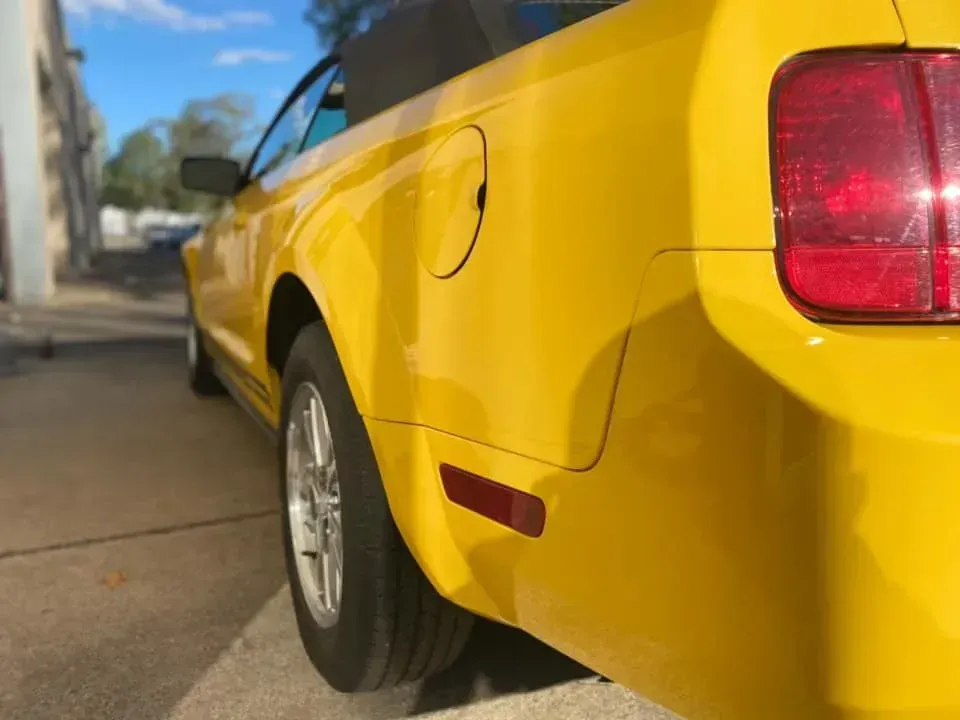 Yellow convertible Mustang, driver-side rear view. Shows tail light, wheel, gas door, and side marker. Sunny outdoor setting.