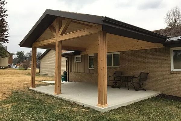 Covered patio with wooden posts and roof, concrete floor, against a brick house. Chairs and a table are on the patio.