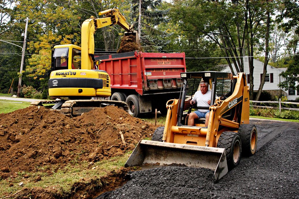 Construction crew with excavator and skid-steer loading dirt beside a red dump truck on a gravel road.