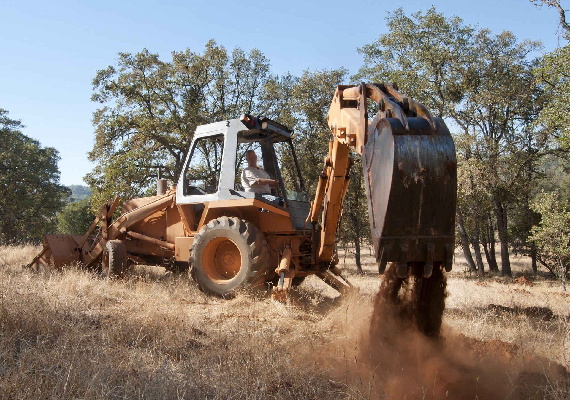Orange backhoe loader digging dry soil in a field, kicking up dust near trees.