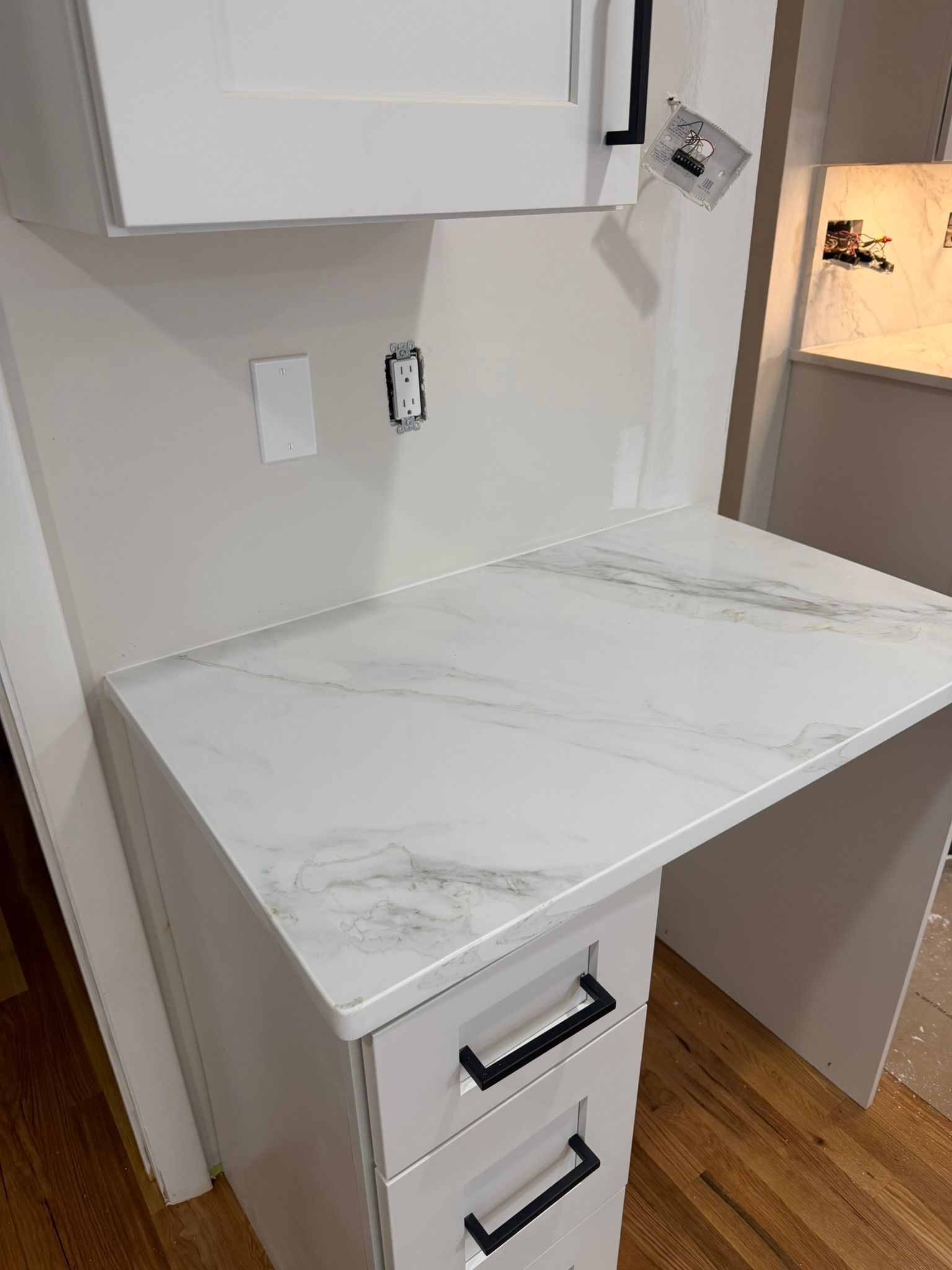 White built-in desk with marble-look top, drawers, and wall cabinets in a small room with hardwood floor