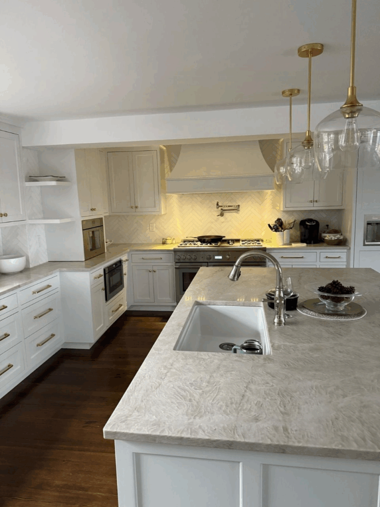 Bright white kitchen with marble island, brass pendant lights, and stainless appliances.