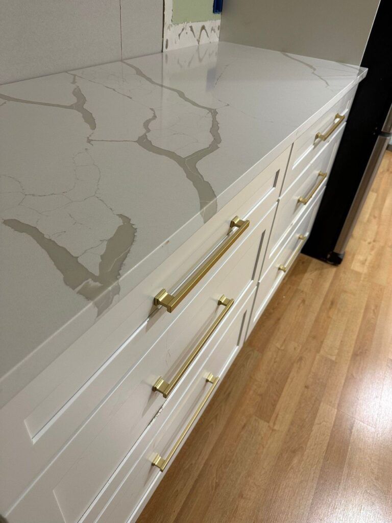 White kitchen counter with marble-like surface above white drawers and brass handles, next to wood flooring
