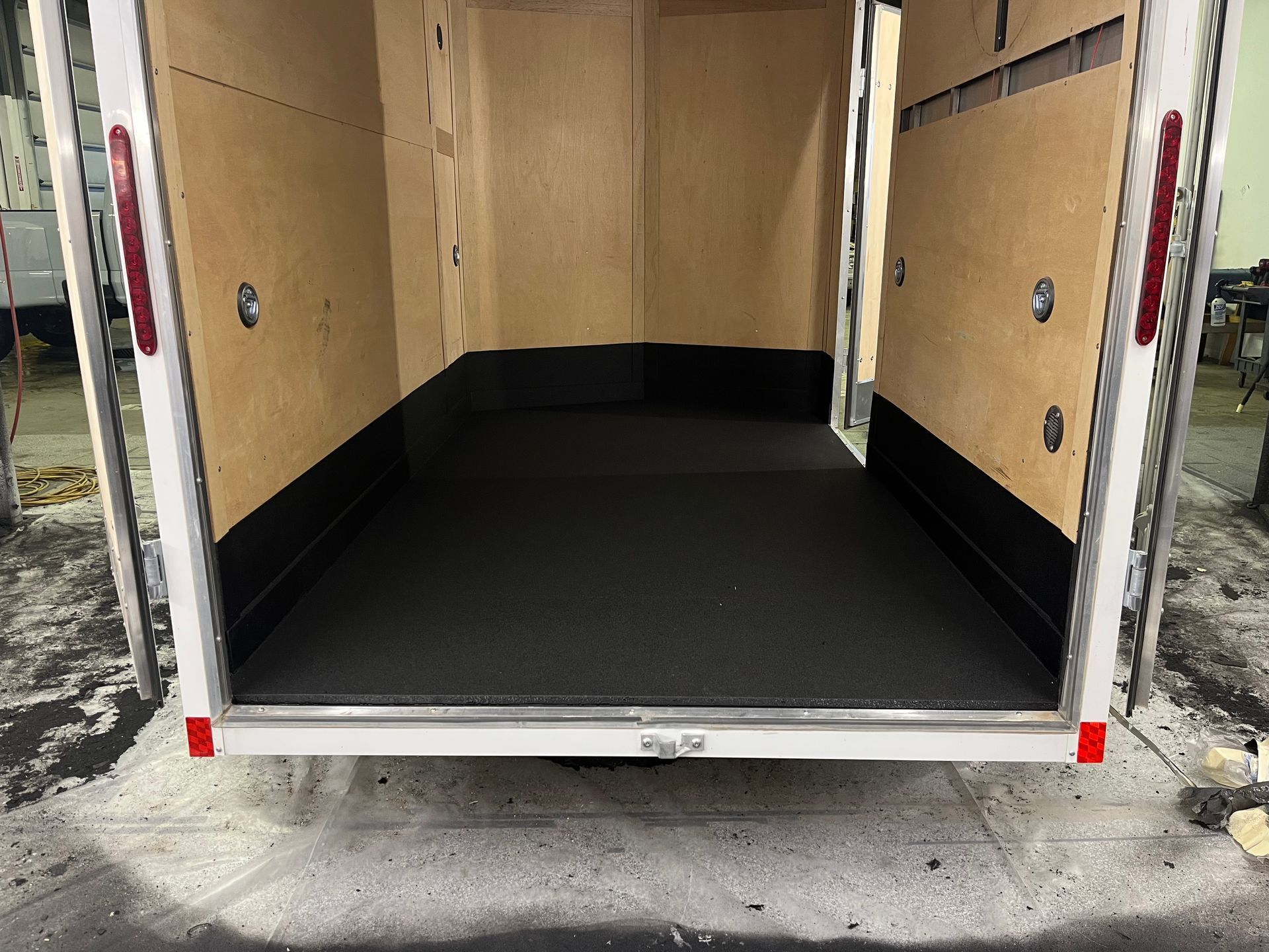 Empty cargo trailer interior with black floor and tan walls, rear door open to a driveway.