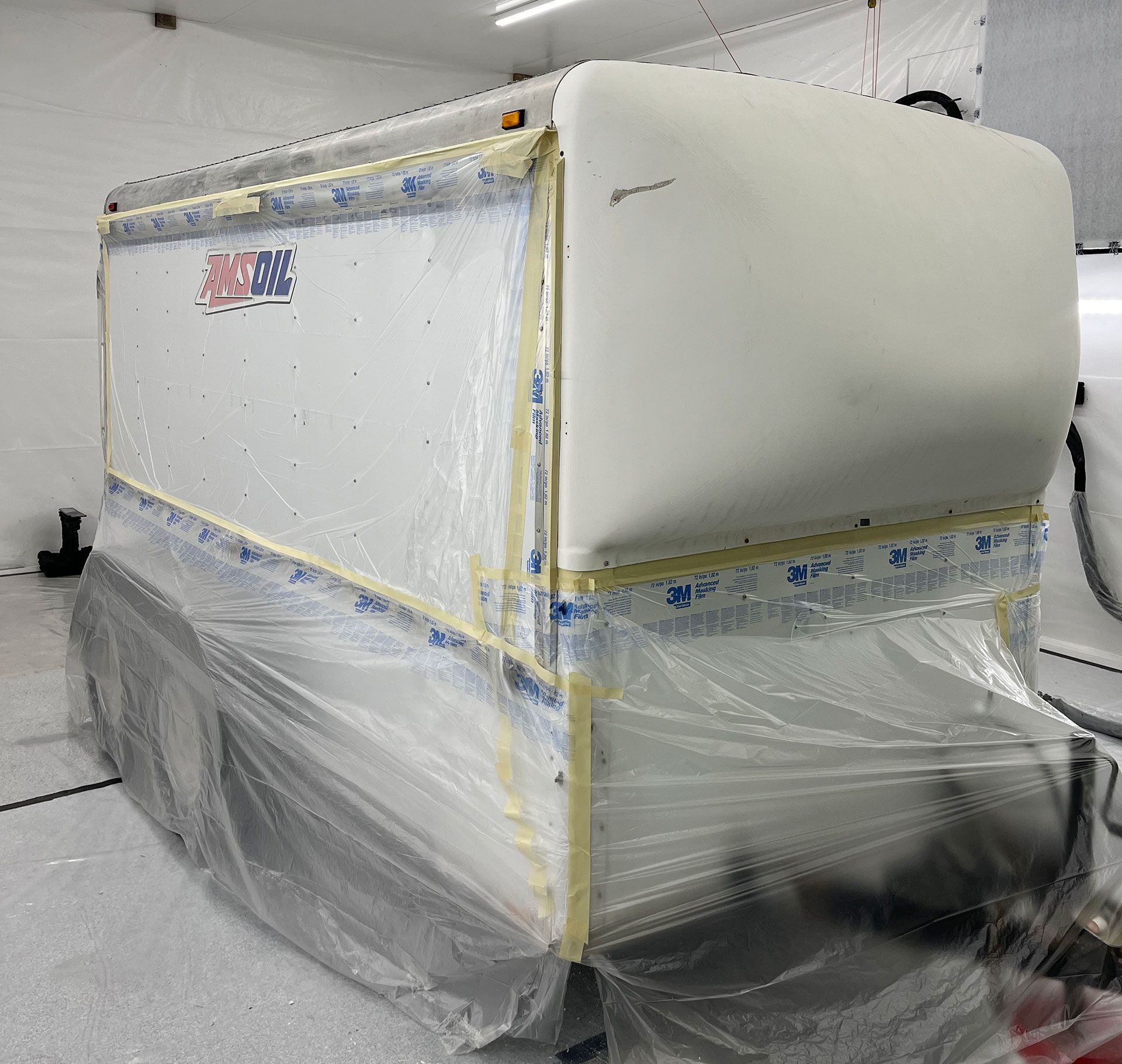 A large trailer masked with plastic sheeting and tape, prepared for paint in a workshop.