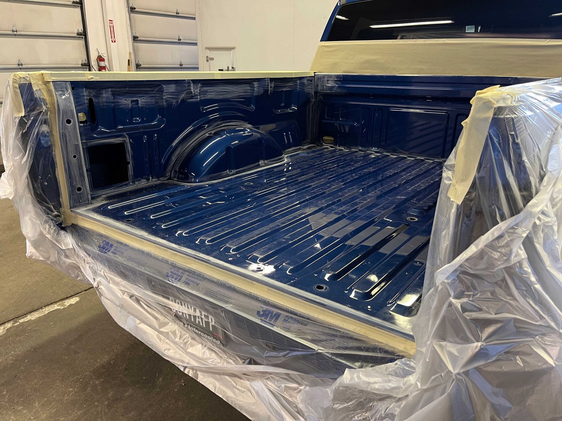 A blue truck bed being prepared for a spray-on bedliner, masked off with tape and plastic sheets in a workshop.