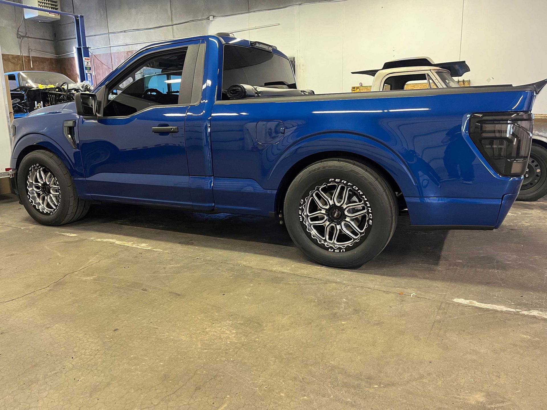A blue single-cab Ford F-150 pickup truck with custom silver and black wheels parked inside a garage.