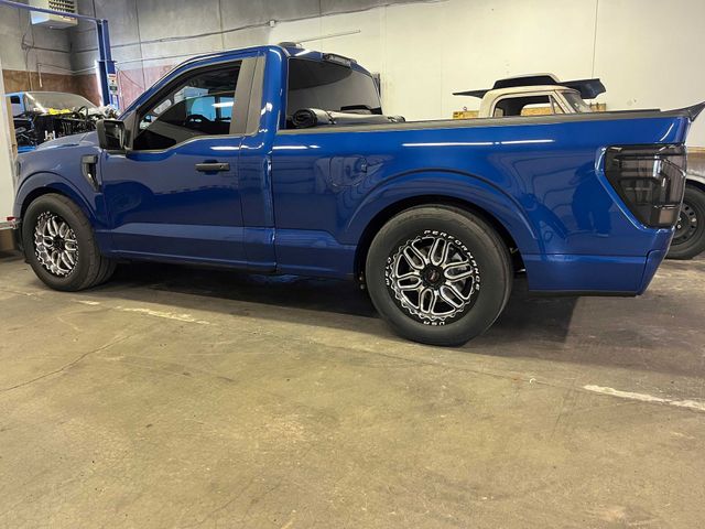A blue single-cab Ford F-150 pickup truck with custom silver and black wheels parked inside a garage.