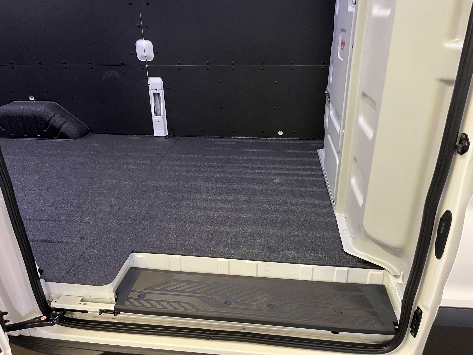 Open side door of a cargo van showing dark gray floor matting, black wall panels, and a plastic entrance step.