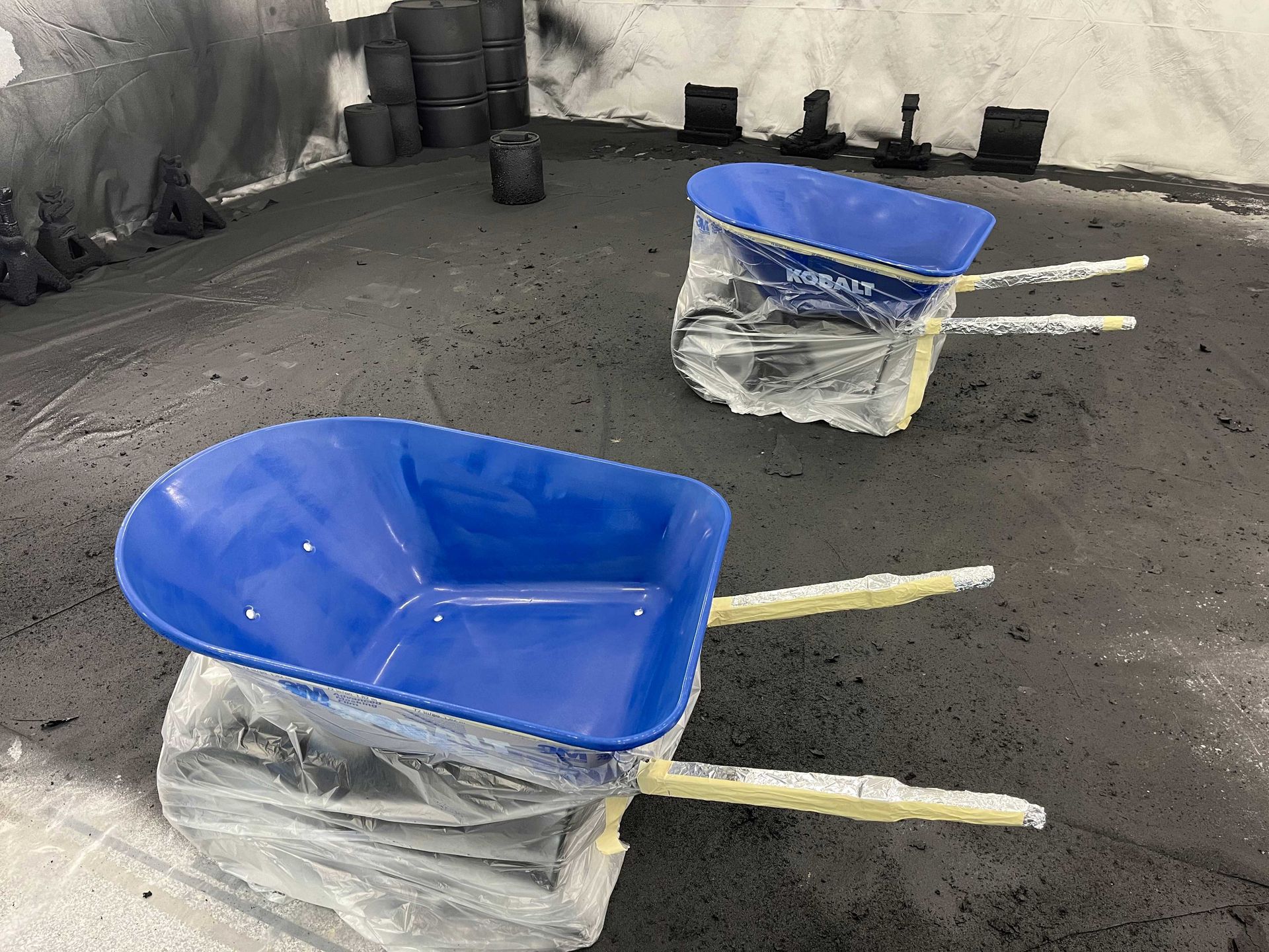 Two blue wheelbarrow tubs wrapped in plastic sit on a concrete floor in a workshop, surrounded by black paint overspray.