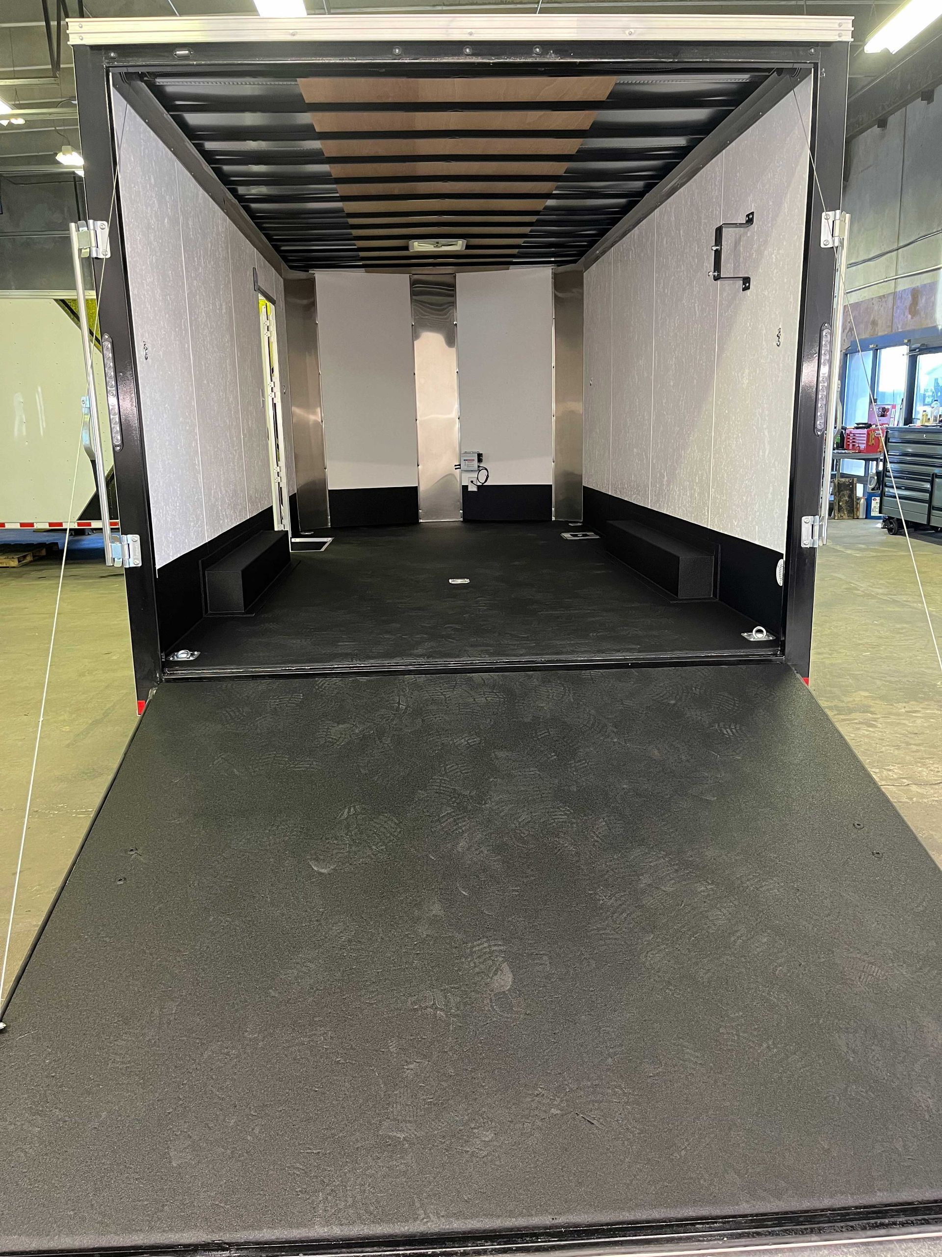 Empty interior of a trailer with black flooring, white walls, and a lowered rear ramp door in an indoor warehouse.