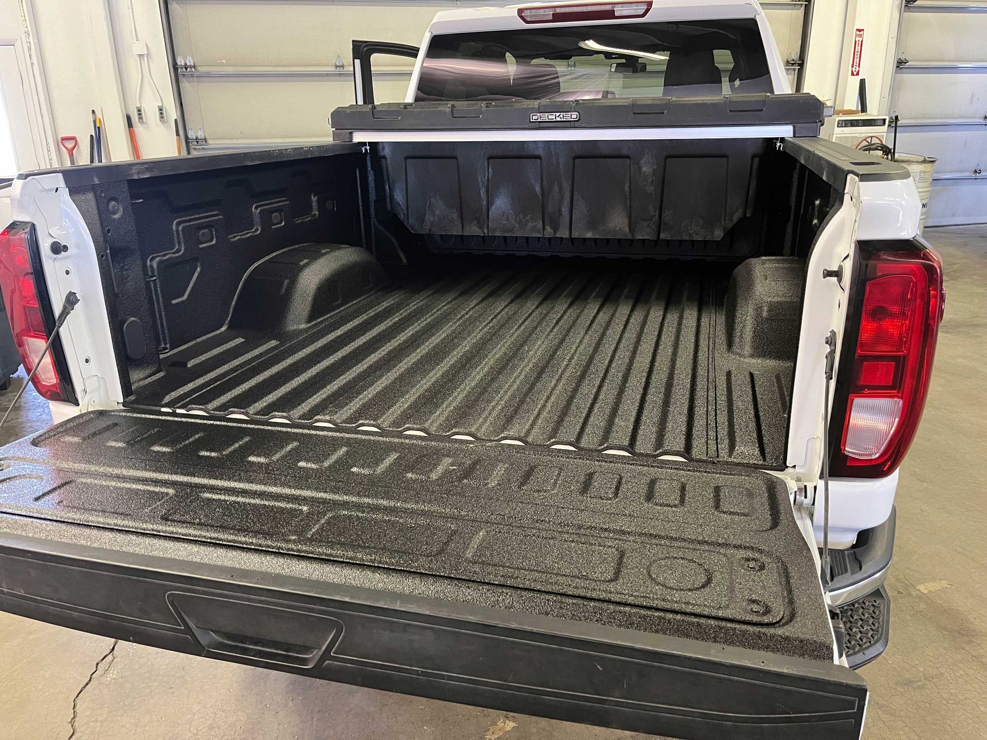 Open truck bed with a black protective spray-on liner, viewed from the rear of a white pickup truck in a garage.