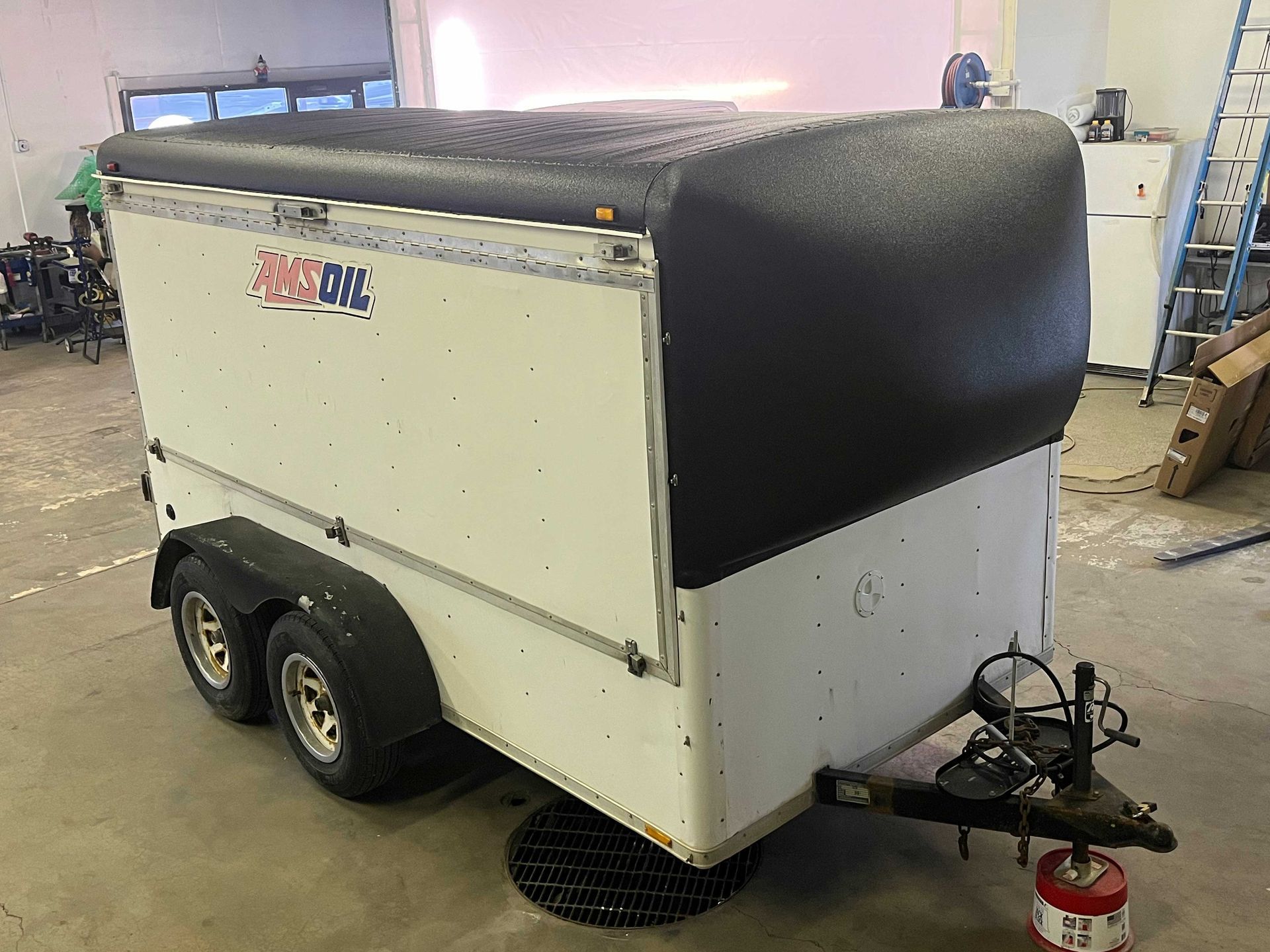 A small, white, tandem-axle enclosed utility trailer with a black, textured aerodynamic nose cone parked in a workshop.